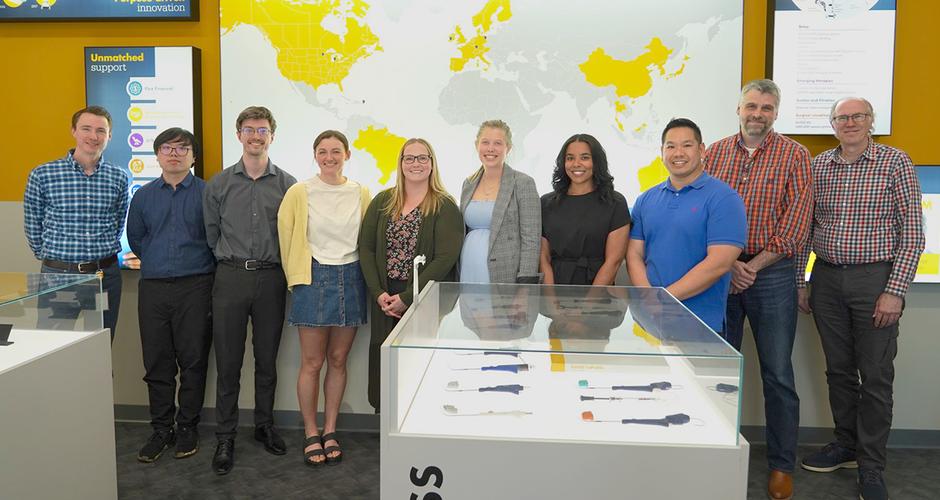 Group of people standing among display cases and against a wall of display screens. Once screen is of a map with some countries filled in with yellow. 